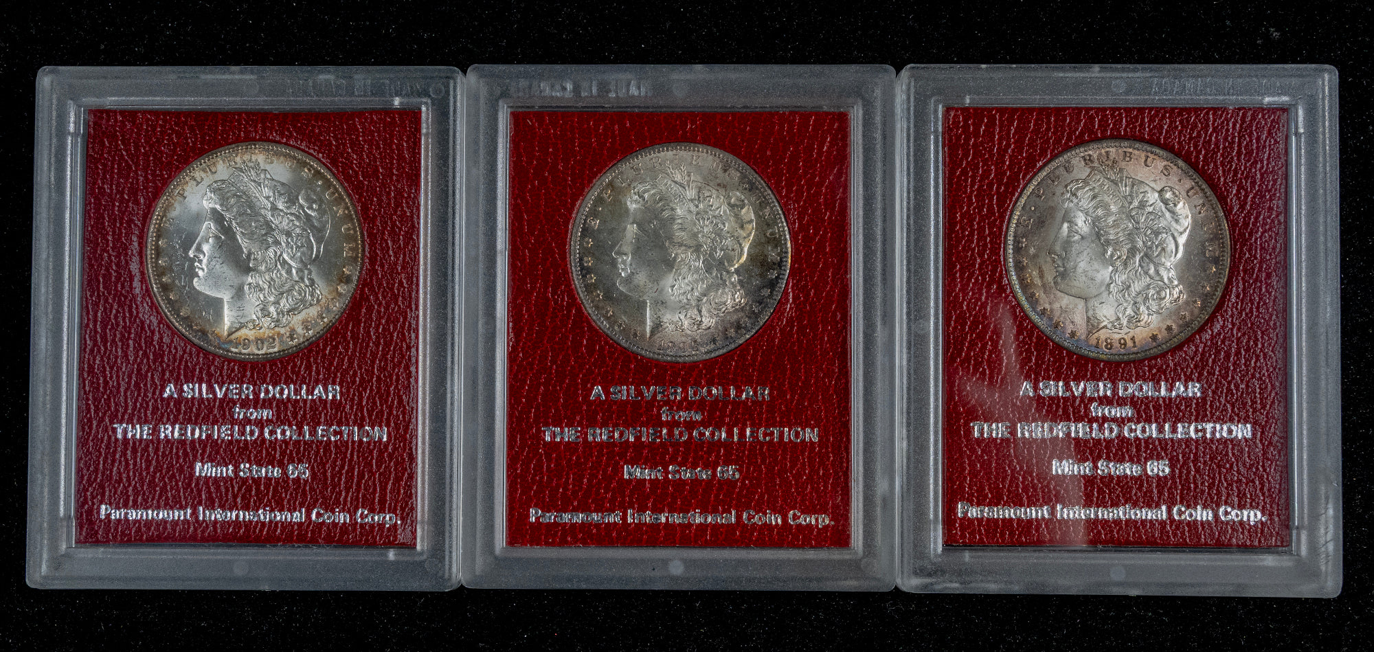 Three silver dollar coins in clear protective cases with red backing cards, arranged side by side on a black background. The cards read 'A SILVER DOLLAR From THE REINFOLD COLLECTION' and show the Permanent International Coin Corp.
