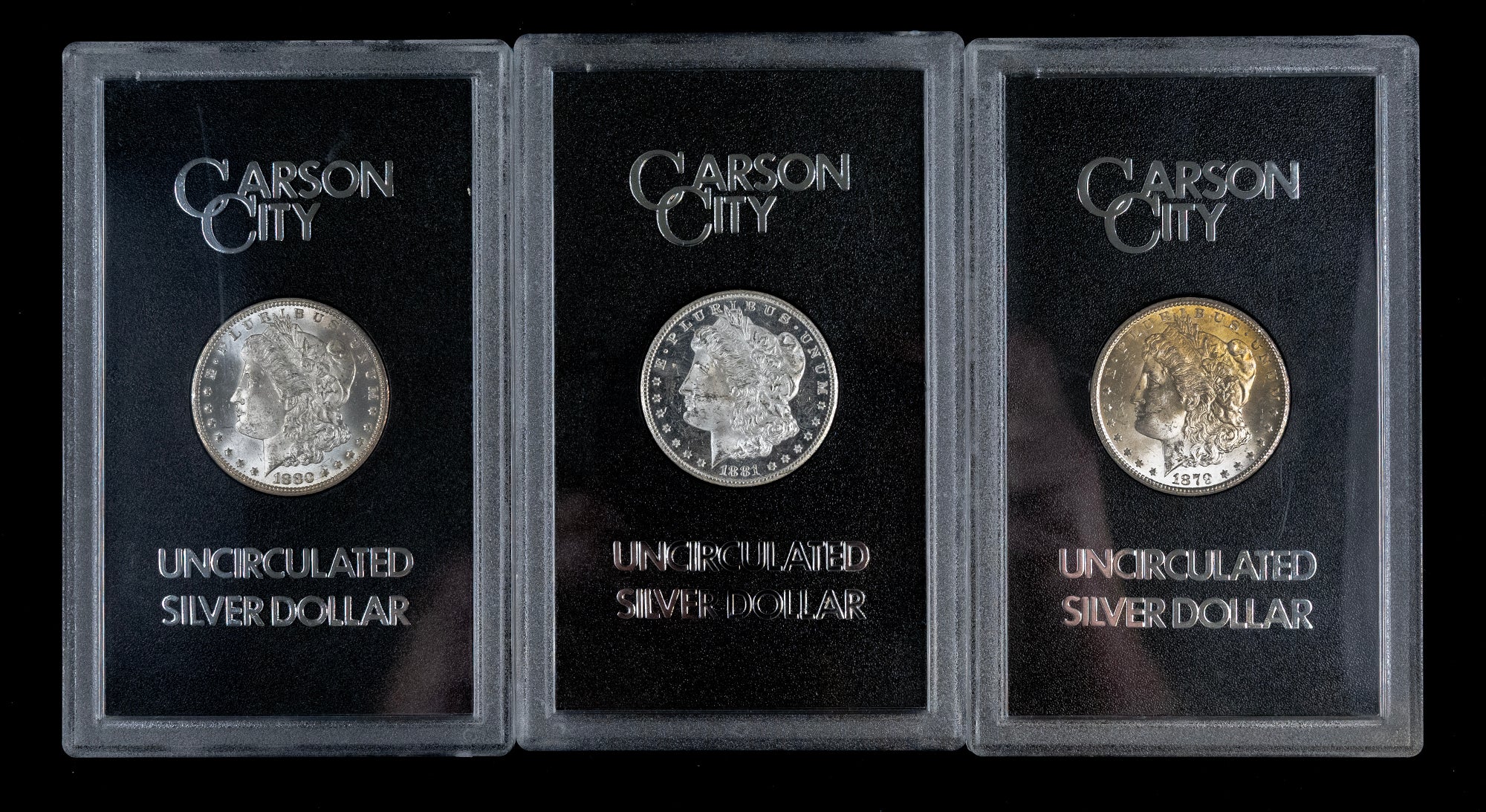 Three Carson City Uncirculated Silver Dollar coins in clear protective plastic cases with black backing cards, shown side by side on a black background.