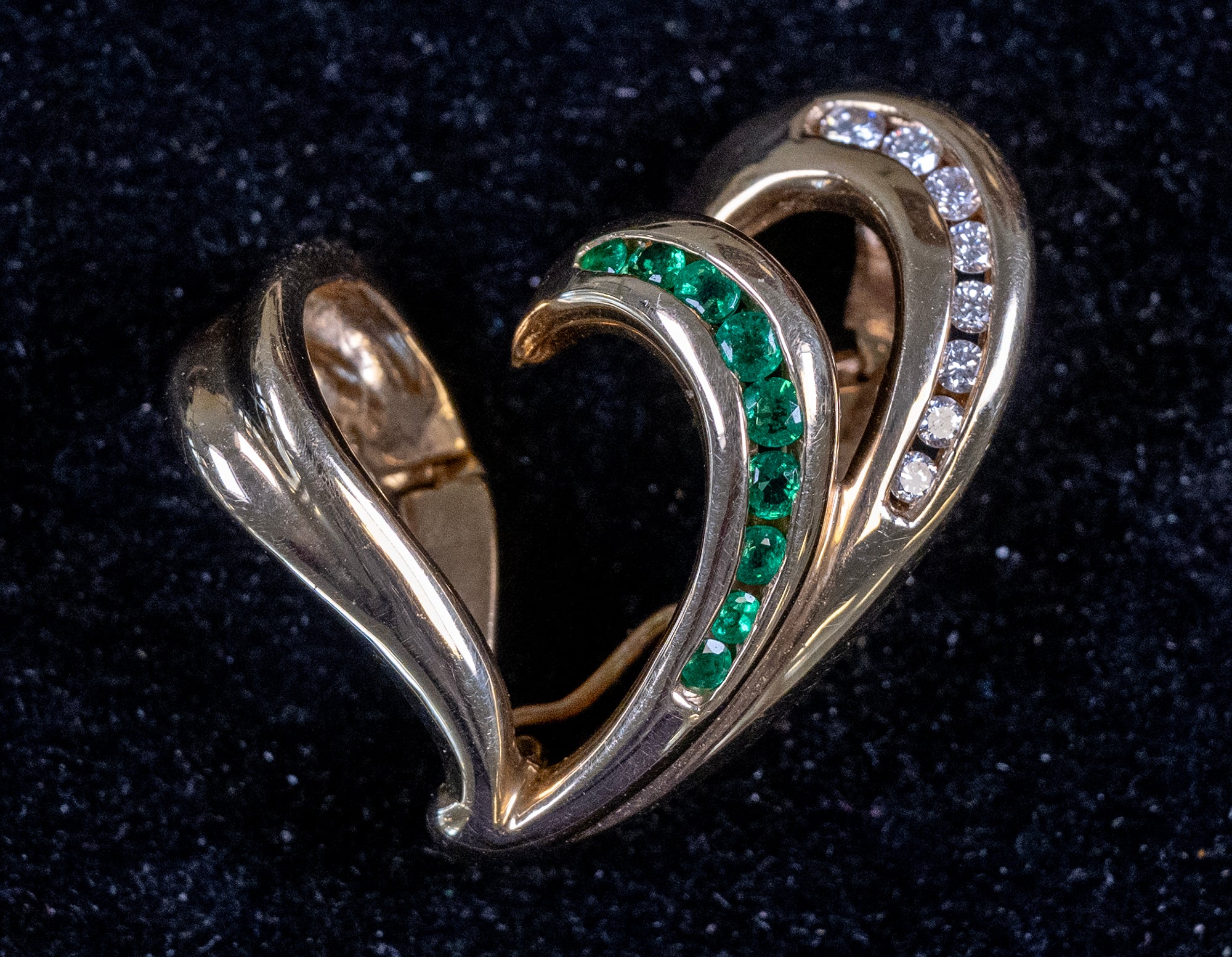 A gold heart-shaped ring brooch featuring a curved row of green emerald-cut stones and a row of clear round-cut stones on the right side, displayed on a dark textured background.