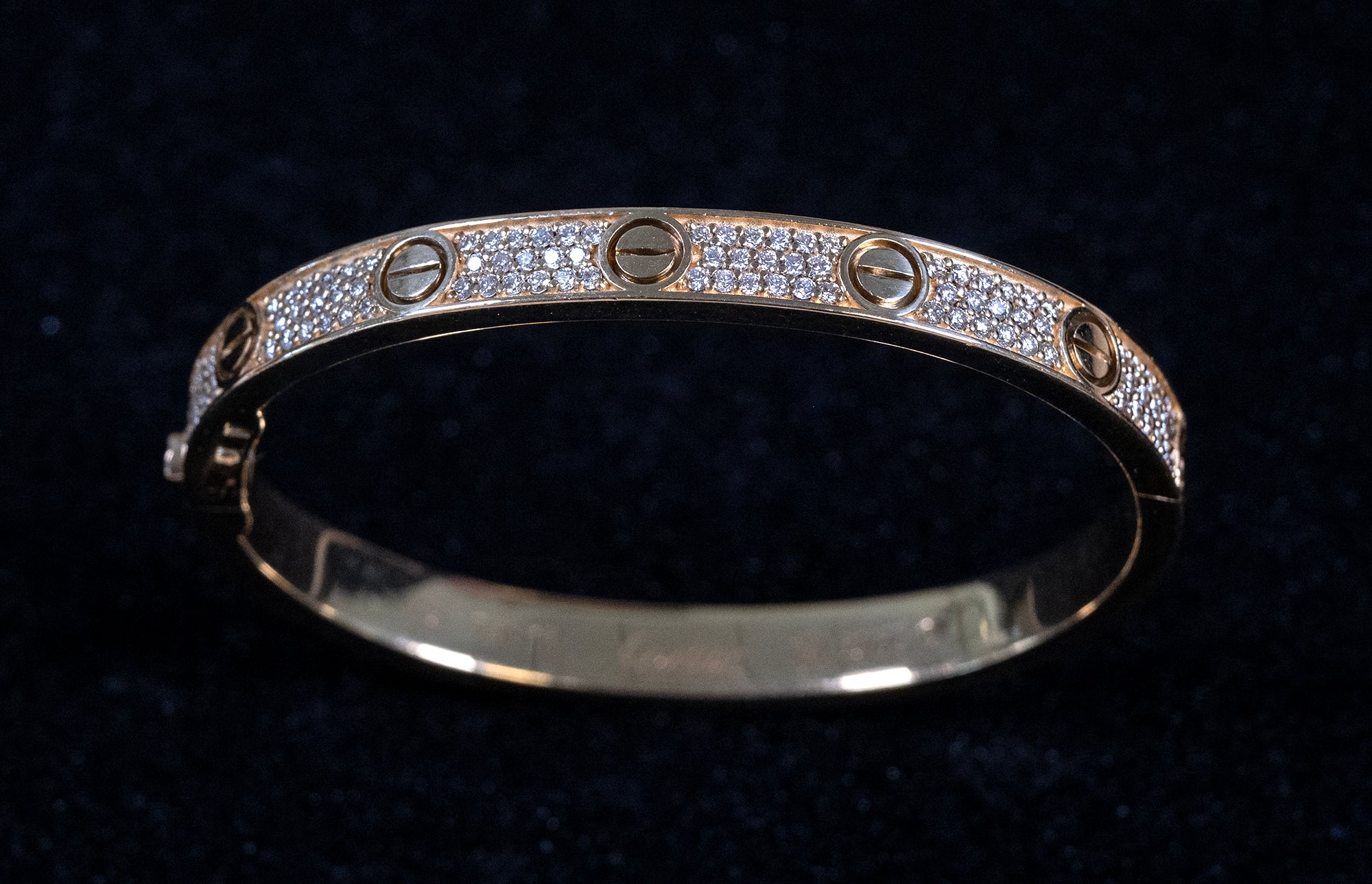 A gold bangle bracelet with a pavé diamond band and screw-head details, displayed on a dark velvet background.