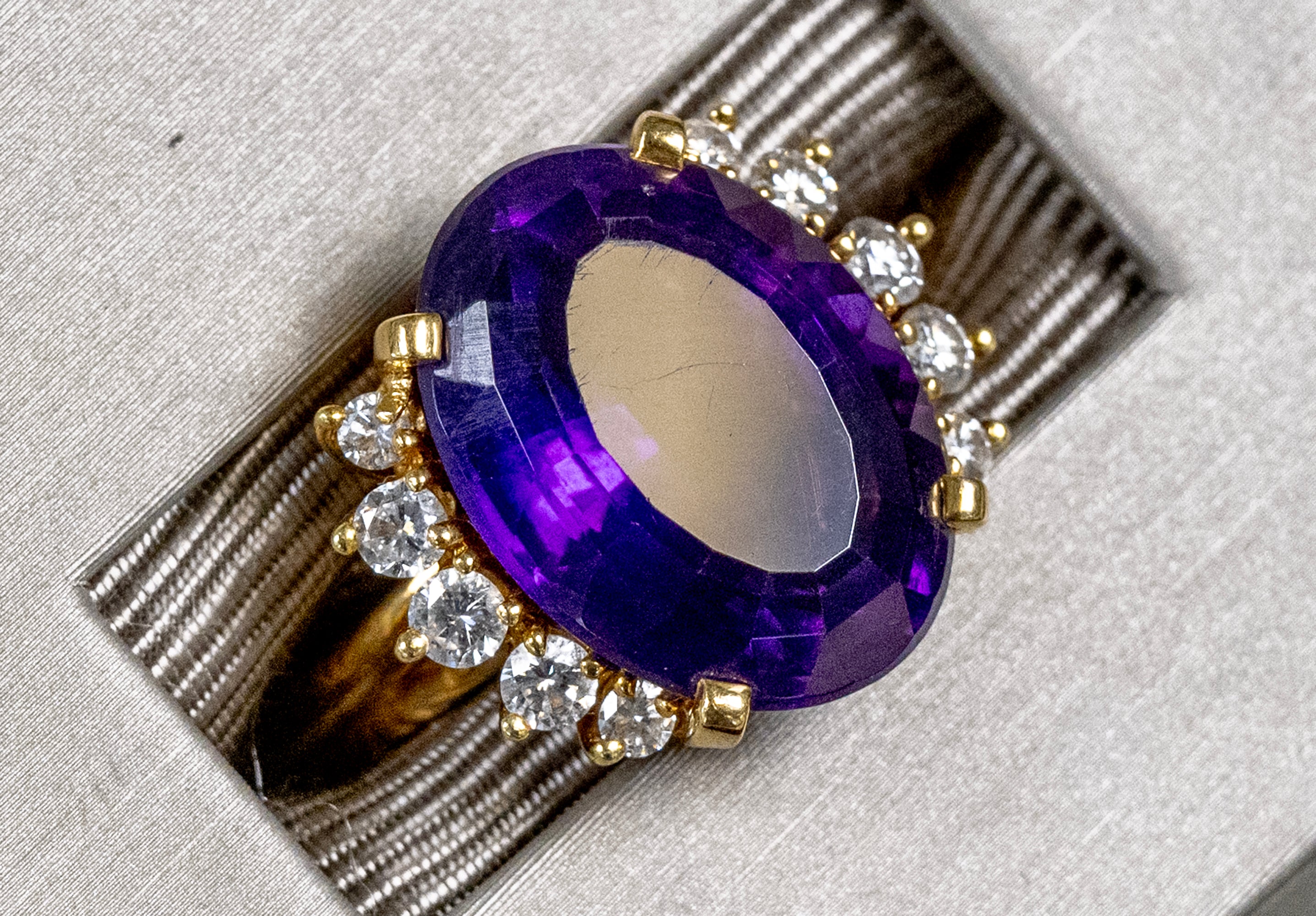 An oval-cut purple amethyst ring with a gold setting and small clear accent diamonds, displayed inside a textured gray jewelry box.