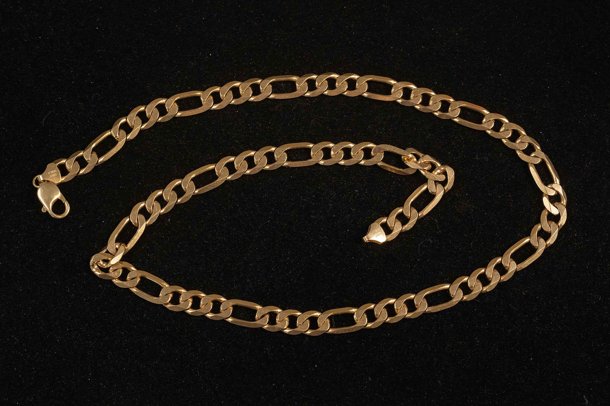 A gold Figaro chain necklace with a lobster clasp, displayed in a loose coil on a black background.