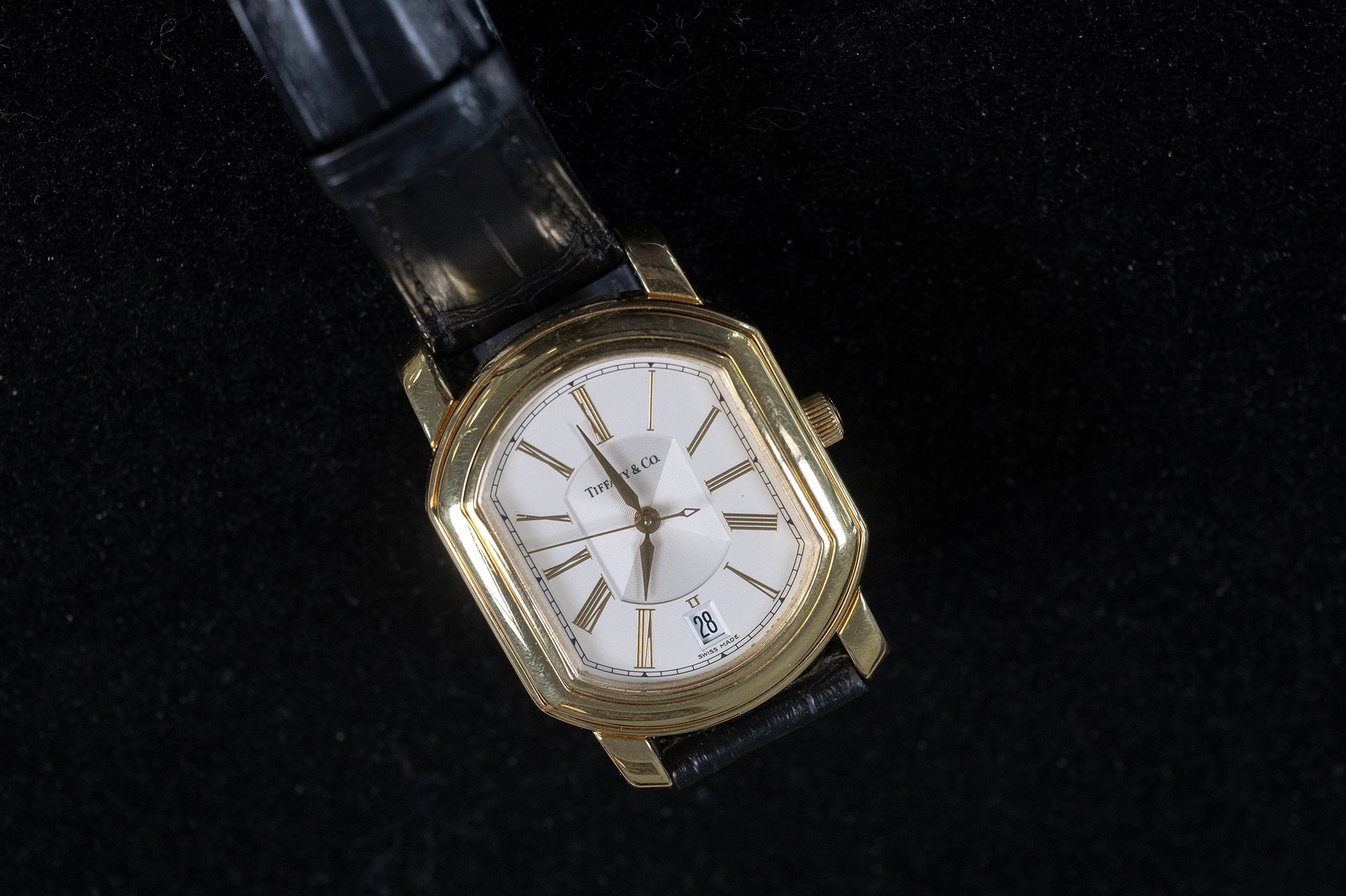 A vintage Tiffany wristwatch with a gold-tone octagonal case, white dial, and black leather strap, photographed on a black textured background.