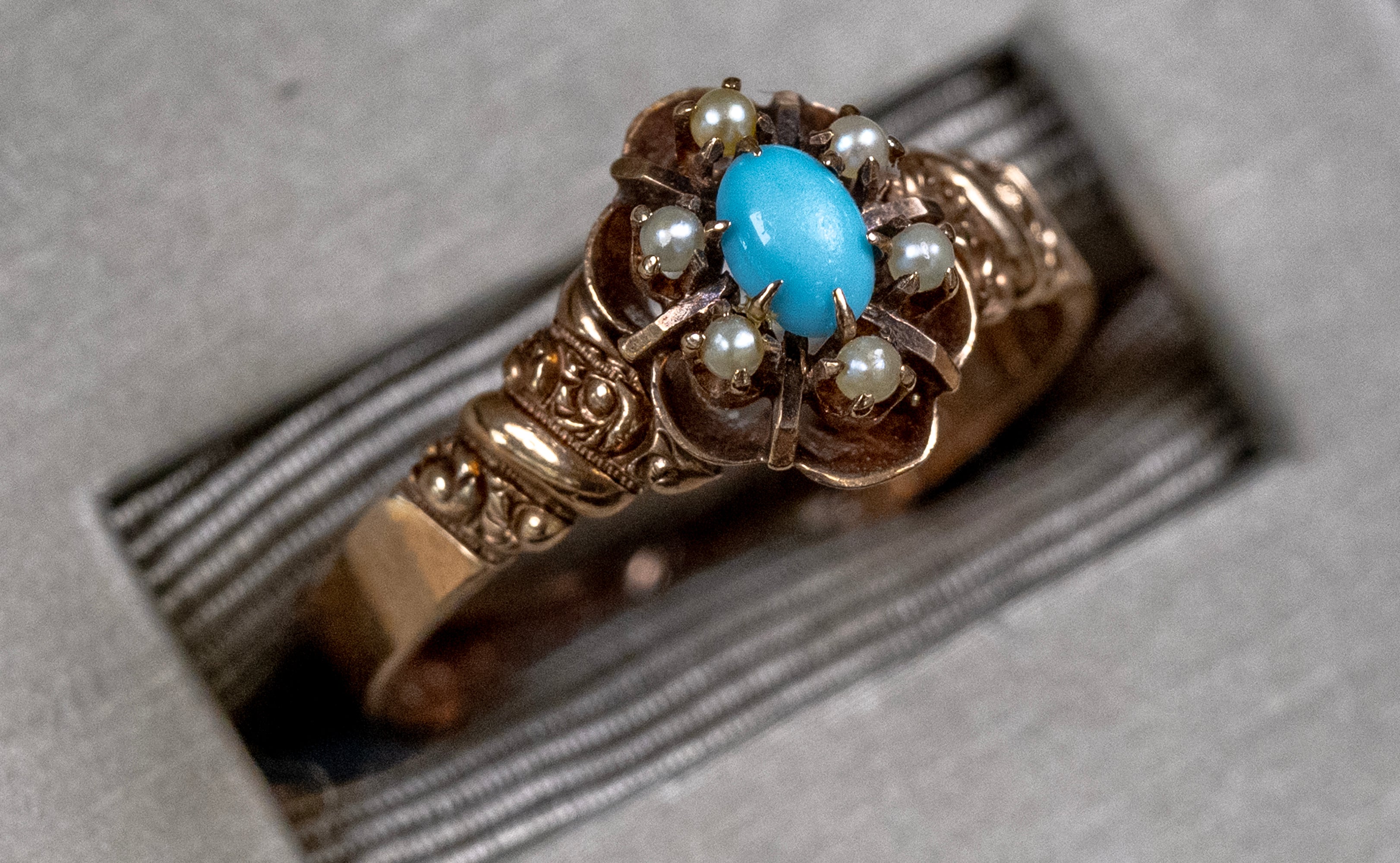 An ornate vintage-style gold ring featuring a central oval turquoise cabochon and surrounding small white pearls, displayed on a textured gray surface.